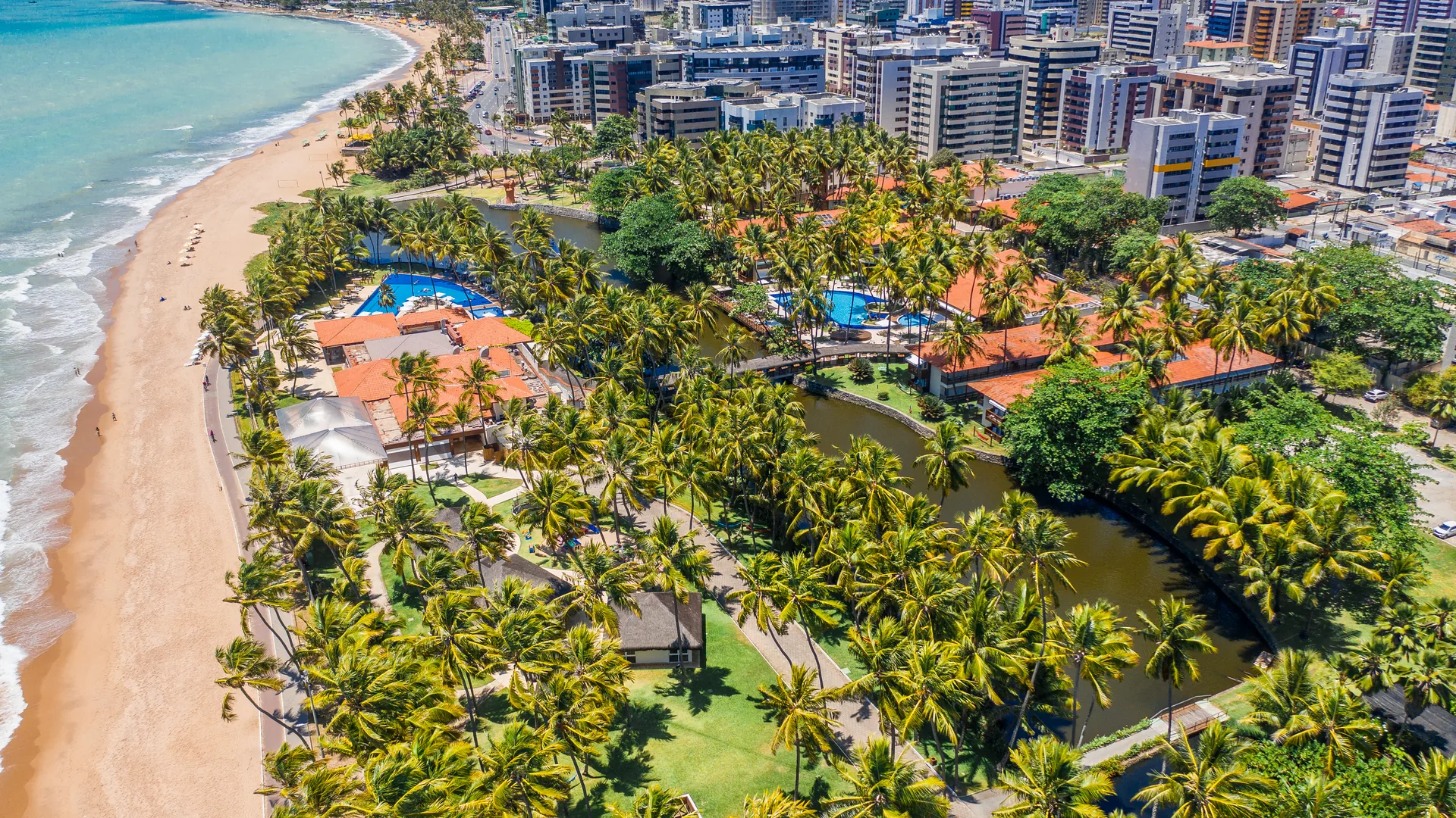 Vista aérea do resort, com as piscinas entre coqueiros e a faixa de areia da orla da Jatiúca ao lado